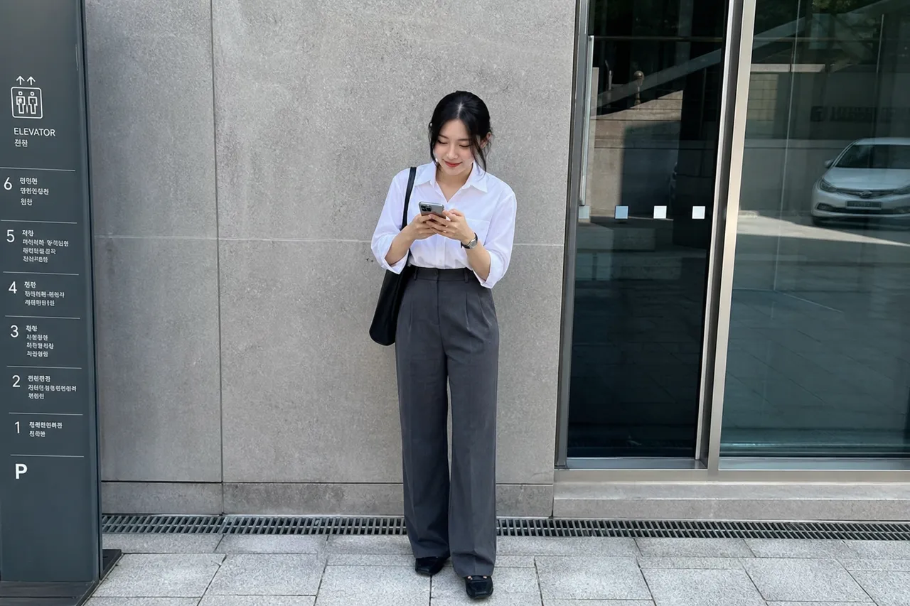 A full-body shot of a Korean woman wearing high-waisted charcoal slacks and a tucked-in white shirt, creating a 3 to 7 body ratio, long leg line effect, urban office background