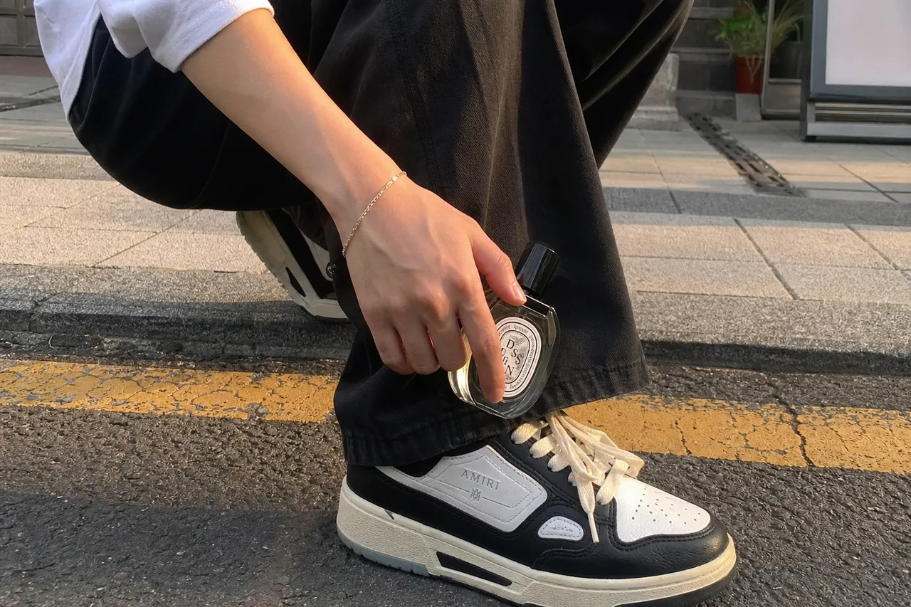 A close-up shot of a person's legs wearing wide-fit pants and trendy sneakers, with a hand holding a perfume bottle near the ankle, urban street style, soft afternoon sunlight
